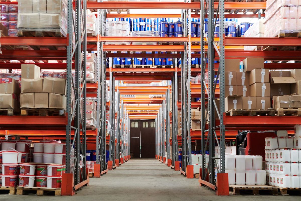 Warehouse aisle with tall orange pallet racking stocked with cartons and drums